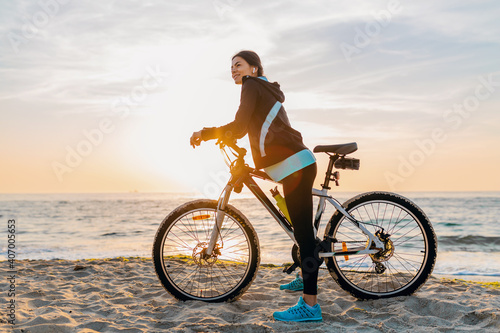young attractive slim woman riding bicycle, sport on morning sunrise summer beach in sports fitness wear, active healthy lifestyle, smiling happy having fun