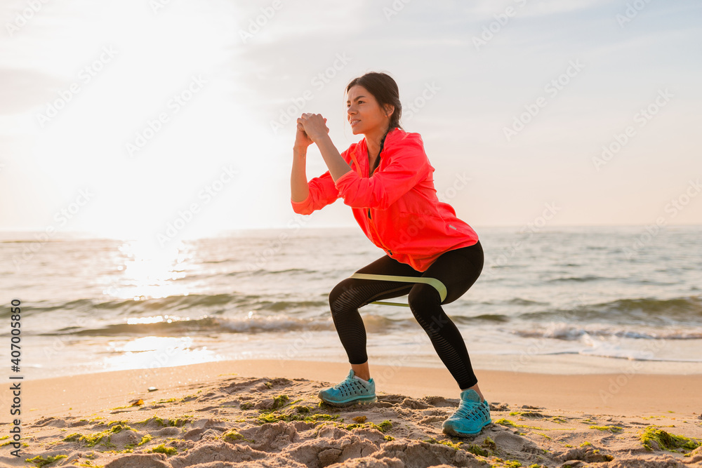 young attractive woman doing sport exercises in morning sunrise on sea beach, healthy lifestyle, listening to music on earphones, wearing pink windbreaker jacket, making stretching in rubber band young attractive woman doing sport exercises in morning sunrise on sea beach, healthy lifestyle, listening to music on earphones, wearing pink windbreaker jacket, making stretching in rubber band