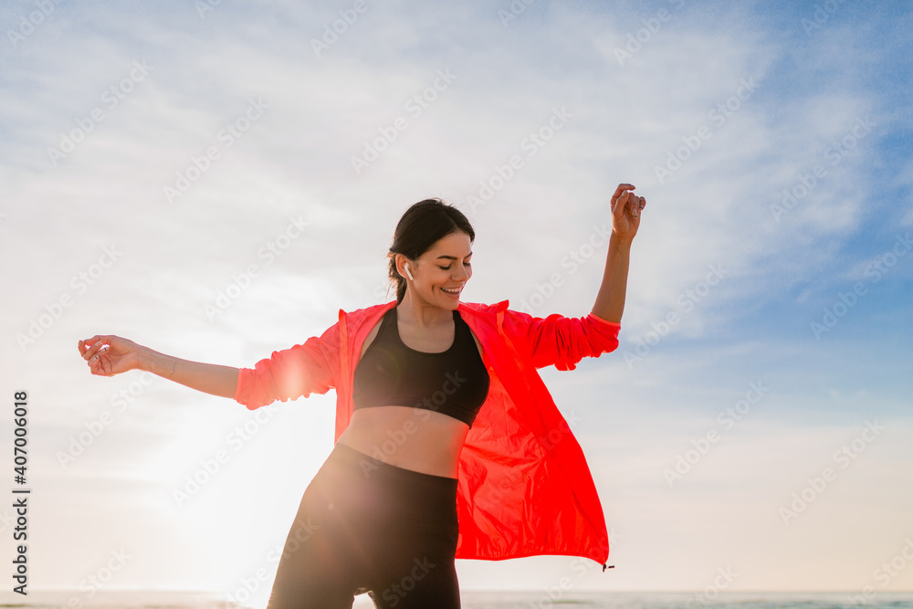 young smiling attractive slim woman doing sports in morning sunrise dancing on sea beach in sports wear, healthy lifestyle, listening to music on earphones, wearing pink windbreaker jacket, having fun young smiling attractive slim woman doing sports in morning sunrise dancing on sea beach in sports wear, healthy lifestyle, listening to music on earphones, wearing pink windbreaker jacket, having fun