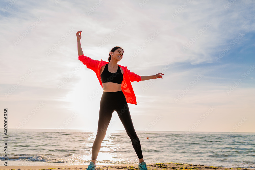 young smiling attractive slim woman doing sports in morning sunrise jumping on sea beach in sports wear, healthy lifestyle, listening to music on earphones, wearing pink windbreaker jacket, having fun
