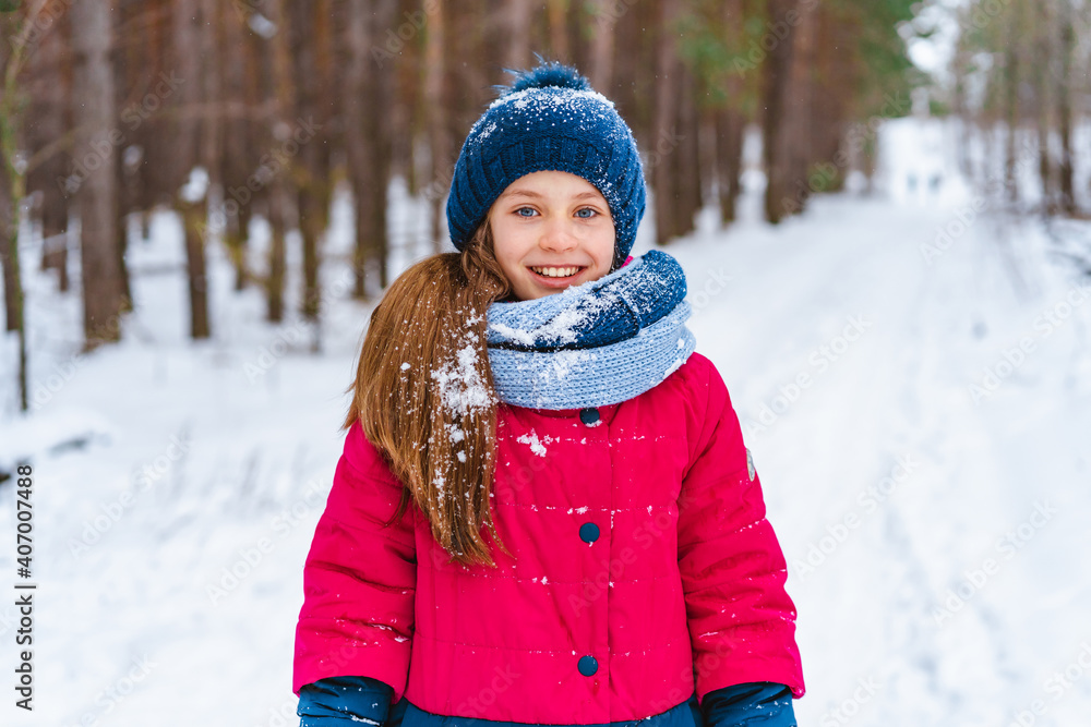 Portrait of a little happy girl in winter in a snow-covered hat who walks in the winter forest