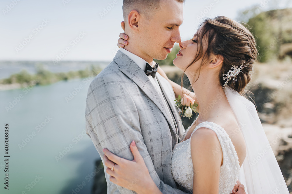A young groom in a gray tailcoat with a black bow tie and a beautiful bride in a white dress are hugging on the background of a cliff, rocks and a river. Wedding portrait of newlyweds. A young groom in a gray tailcoat with a black bow tie and a beautiful bride in a white dress are hugging on the background of a cliff, rocks and a river. Wedding portrait of newlyweds.