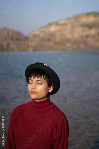 Portrait of an aesthetic looking girl wearing red sweater or sweatshirt with a black hat captured near a lake with a beautiful scenery in the background. © Yogendra