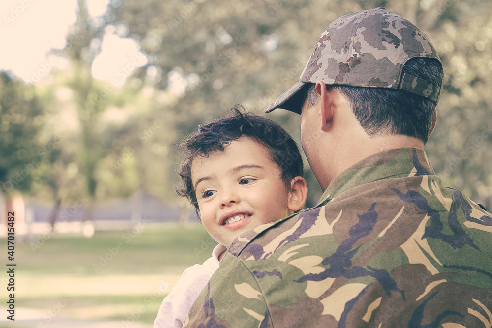 Back view of Caucasian man holding kid and wearing army uniform. Cheerful little boy sitting on father hands, hugging dad and smiling happily. Family reunion, fatherhood and returning home concept