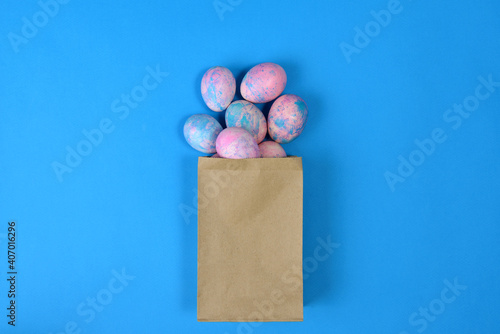 Easter eggs painted in blue and pink lie in a brown paper bag on a blue background, top view. Several colorful Easter eggs are spilled out of paper craft packaging