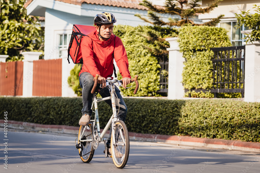 A courier in red uniform with delivery box on back riding bicycle and looking on cellphone to check addres to deliver food to customer. Courier on bicycle delivering food in city. A courier in red uniform with delivery box on back riding bicycle and looking on cellphone to check addres to deliver food to customer. Courier on bicycle delivering food in city.