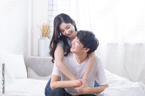 Young Asian couple wife hugging husband from behind and look at each other and smiling together bed room. Romantic moment.