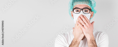 Doctor surgeon in white medical mask and a medical cap stands over white background with surprise,covers his mouth with the hands. Сoncept of covid 19, flu and seasonal cold.