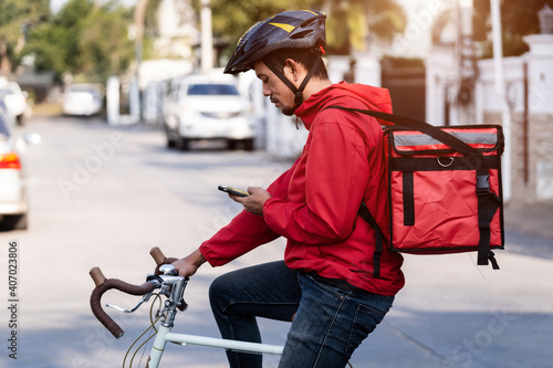 A courier in red uniform with delivery box on back riding bicycle and looking on cellphone to check addres to deliver food to customer. Courier on bicycle delivering food in city. photo