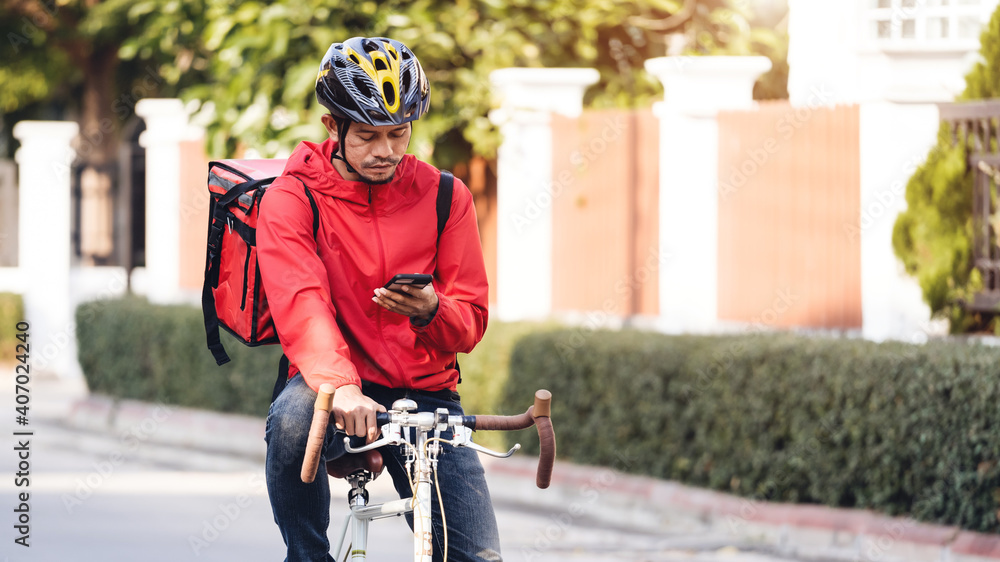 Naklejka premium A courier in red uniform with delivery box on back riding bicycle and looking on cellphone to check addres to deliver food to customer. Courier on bicycle delivering food in city.