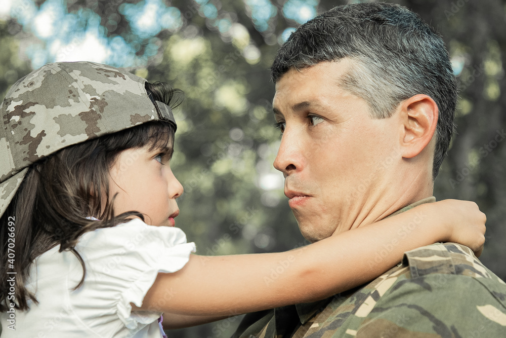 Positive dad in camouflage uniform holding offended daughter in arms, hugging girl outdoors after returning from military mission trip. Closeup shot. Family reunion or returning home concept Positive dad in camouflage uniform holding offended daughter in arms, hugging girl outdoors after returning from military mission trip. Closeup shot. Family reunion or returning home concept