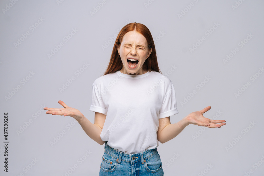 Angry frustrated young woman wearing T-shirt and denim pants screaming out loud on isolated white background. Pretty lady model with red hair emotionally showing facial expressions. Angry frustrated young woman wearing T-shirt and denim pants screaming out loud on isolated white background. Pretty lady model with red hair emotionally showing facial expressions.