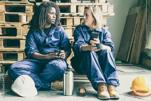 Two diverse female factory worker drinking coffee in warehouse, talking and using cell. Labor or communication concept