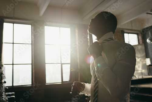 African american man straightens striped tie with hand near face in sun glare in apartment