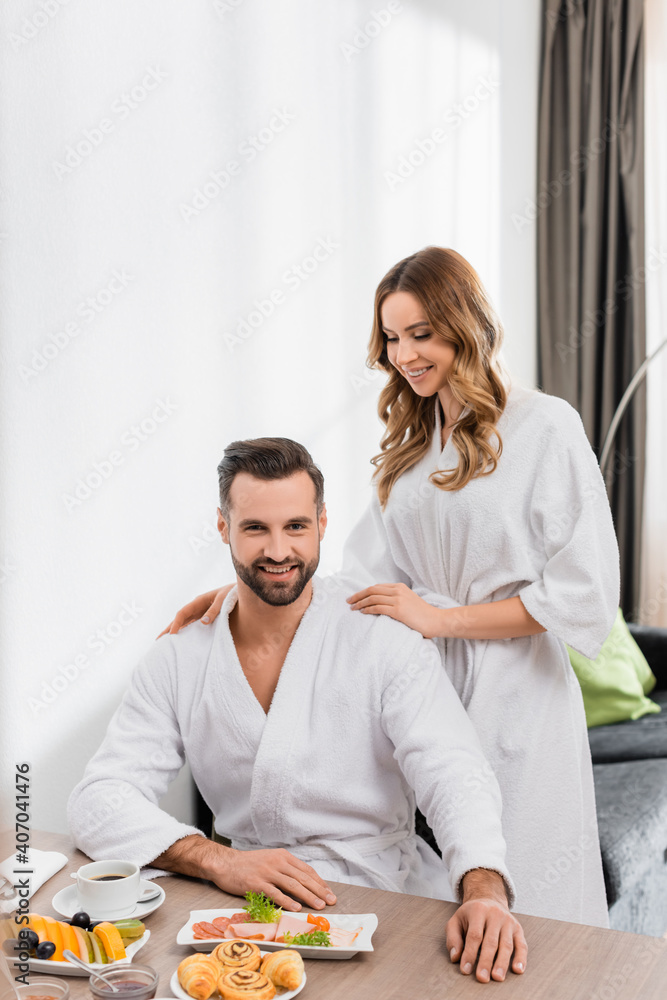 Cheerful woman in bathrobe hugging boyfriend near breakfast with coffee on table in hotel room Cheerful woman in bathrobe hugging boyfriend near breakfast with coffee on table in hotel room