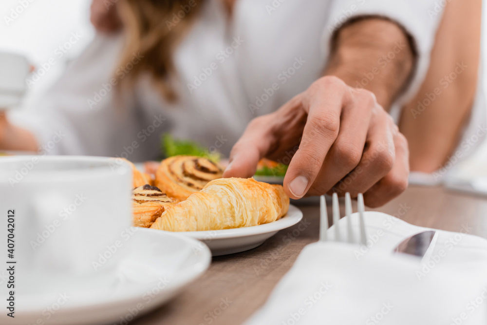 Cropped view of man taking croissant near girlfriend and cup on blurred foreground in hotel Cropped view of man taking croissant near girlfriend and cup on blurred foreground in hotel
