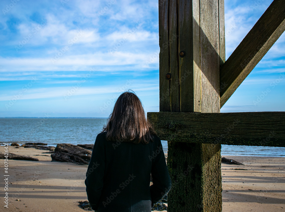 Vendee, France; January 17, 2021: Photo of a young woman looking at the ocean with a long black coat, standing at the foot of the Ile de Noirmoutier boom.