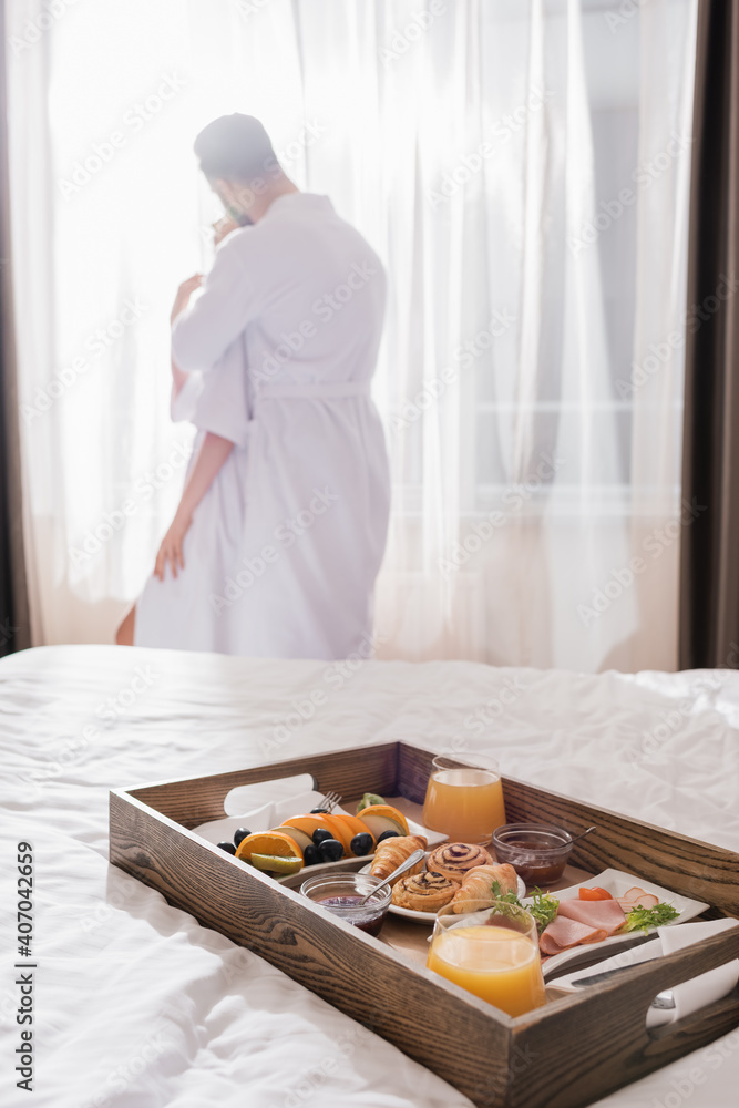 Fototapeta premium Breakfast on wooden tray on bed and couple in bathrobes embracing near window on blurred background in hotel room