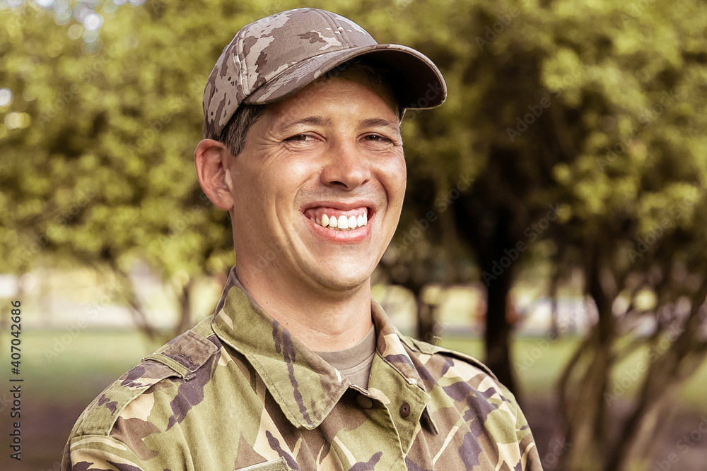 Portrait of happy man in military camouflage uniform standing in park, looking at camera and smiling. Green trees in background. Military man or guard concept