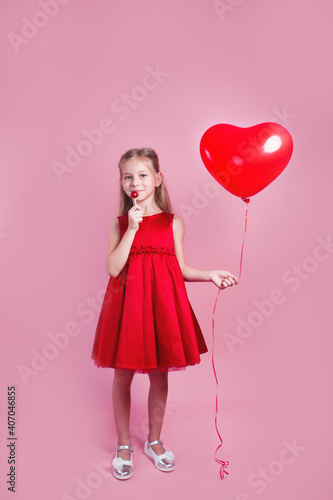 Valentines day. Little girl in red dress with heart shape air balloon on pink background