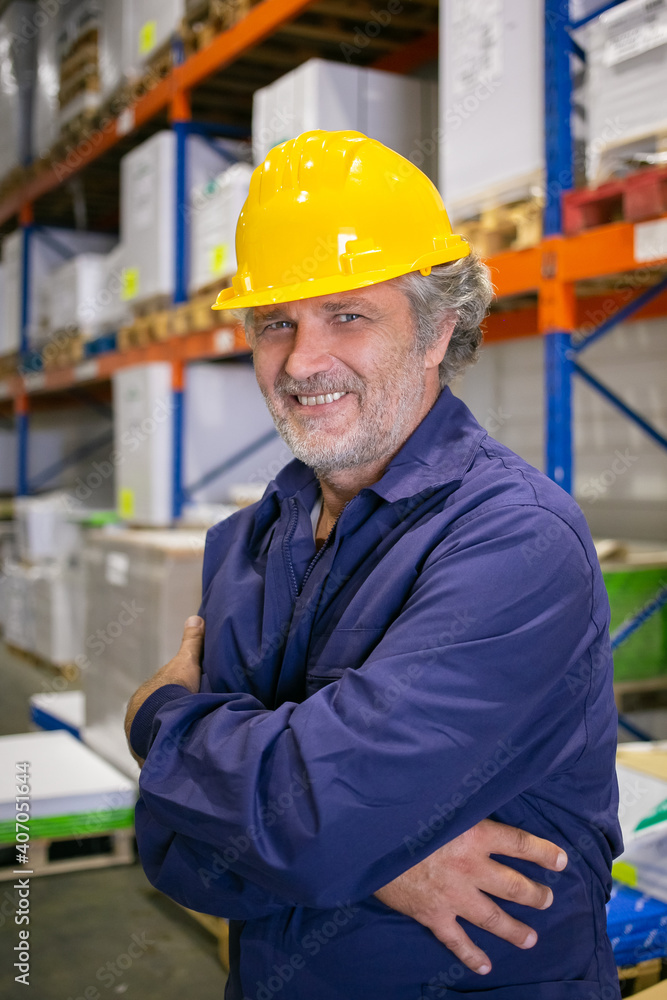 Cheerful grey haired logistic worker in hardhat and uniform standing at shelves in warehouse with arms folded, looking at camera and smiling. Vertical shot. Labor and blue collar portrait concept Cheerful grey haired logistic worker in hardhat and uniform standing at shelves in warehouse with arms folded, looking at camera and smiling. Vertical shot. Labor and blue collar portrait concept