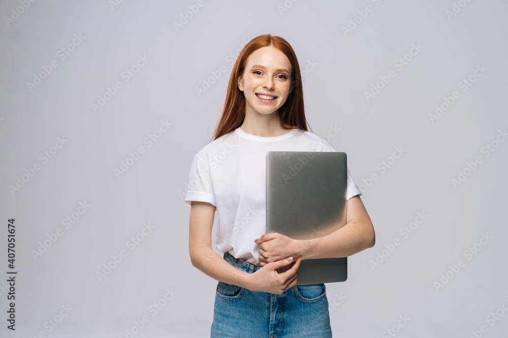 Smiling young woman student holding laptop computer and looking at camera on isolated gray background. Pretty lady model with red hair emotionally showing facial expressions in studio, copy space. Smiling young woman student holding laptop computer and looking at camera on isolated gray background. Pretty lady model with red hair emotionally showing facial expressions in studio, copy space.
