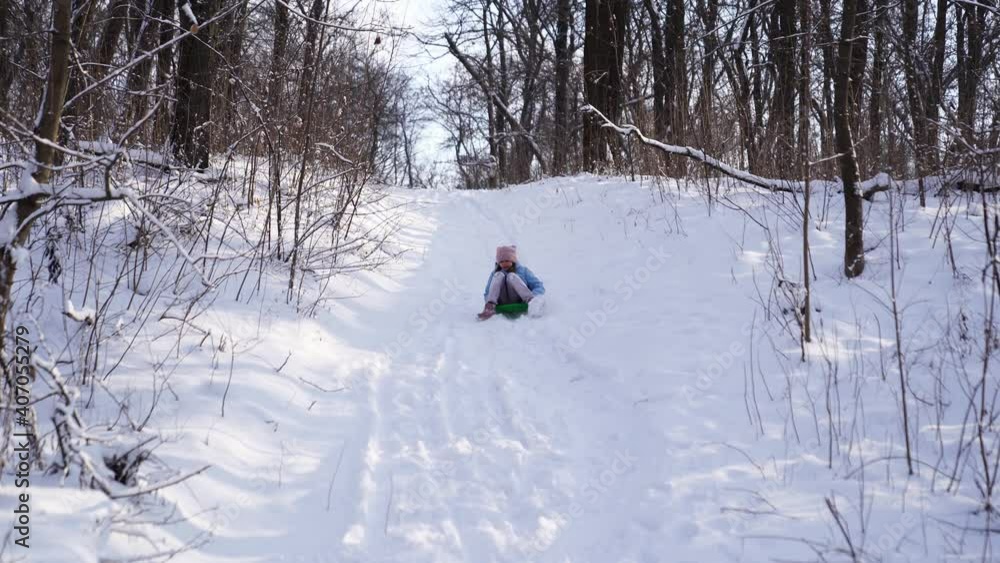 A teenage girl in a pink hat and a blue coat tries to move on a green sledge from a snow slide, but the snow is a lot and it does not slide. winter children's walks and fun. clothes for frosts. A teenage girl in a pink hat and a blue coat tries to move on a green sledge from a snow slide, but the snow is a lot and it does not slide. winter children's walks and fun. clothes for frosts.