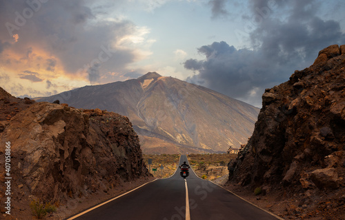 A motorcyclist has passed through a gorge and is driving on the straight and hilly road towards Mount Teide on the island of Tenerife. It is just before sunset in the evening.