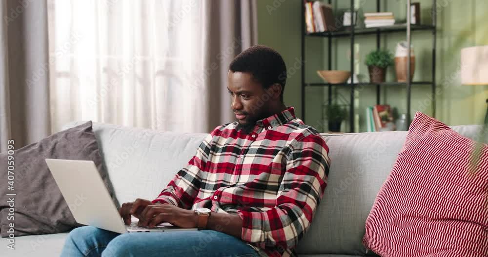 African American young handsome serious man sitting on couch in living room typing on computer working home on laptop with busy face. Freelance work, studying in house, education concept