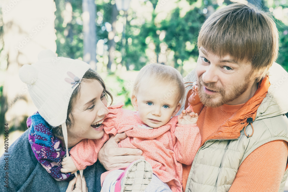 Fototapeta premium Joyful parents and toddler daughter having fun while walking outdoors, posing and laughing. Dad holding kid, mom wearing funny girls hat. Family portrait concept