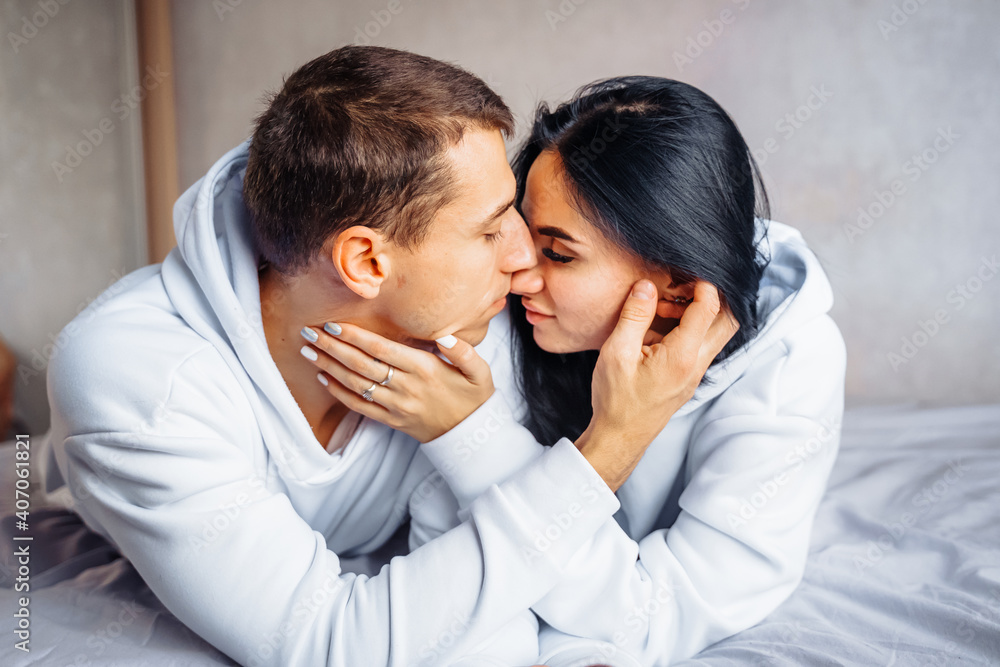 Cute, charming, attractive young Europeans man and woman lie relaxed on the bed. Looking out the window, hug, stroke each other. Foreheads, faces together, kiss. Tenderness at home Cute, charming, attractive young Europeans man and woman lie relaxed on the bed. Looking out the window, hug, stroke each other. Foreheads, faces together, kiss. Tenderness at home