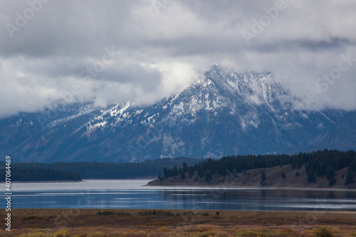 A Teton Mountain with snow and clouds and a lake in the foreground