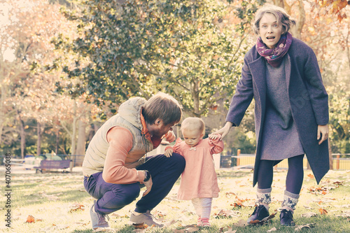 Cheerful family with little girl walking in autumn park on lawn. Blonde mum standing with open mouth and looking forward. Bearded dad squatting and talking with kid. Parenthood and holiday concept