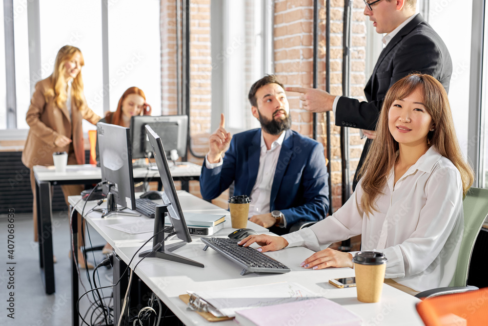 korean woman sitting at desk, at work place, enjoying work with colleagues standing in the background, she looks at camera smiling korean woman sitting at desk, at work place, enjoying work with colleagues standing in the background, she looks at camera smiling