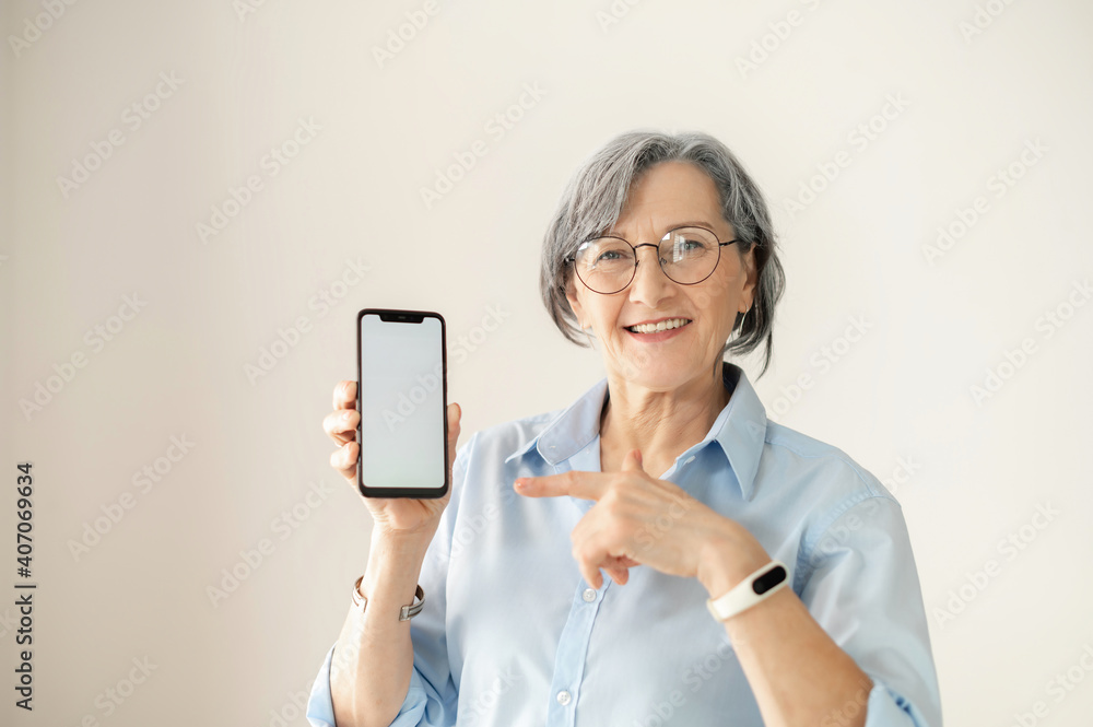 Portrait of a senior elder smiling woman showing or presenting mobile phone application and pointing finger at the smartphone screen on hand, mature businesswoman isolated over gray background Portrait of a senior elder smiling woman showing or presenting mobile phone application and pointing finger at the smartphone screen on hand, mature businesswoman isolated over gray background
