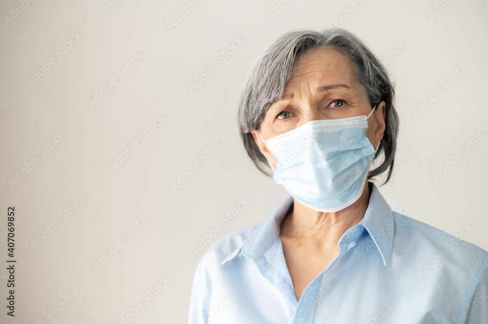 Portrait of a sad senior middle-aged female businesswoman with gray hair wearing a face mask, looking at the camera, headshot of doctor at the workplace, stay safe, stay at home, protect grandparents