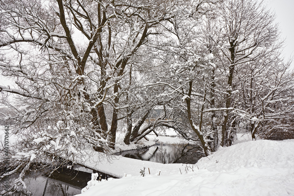 Small river in winter. Rural January scene, bridge over creek. Trees on riverbank covered with snow.