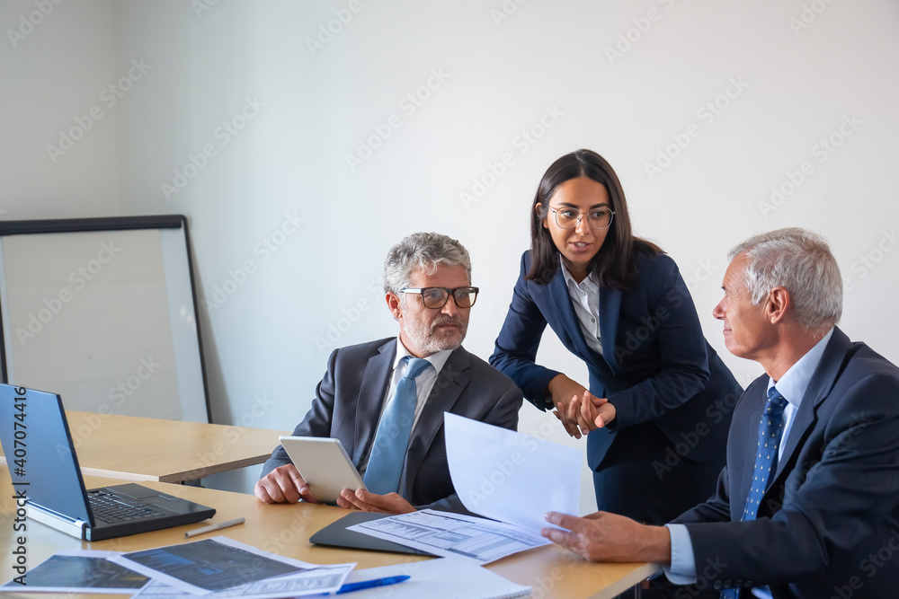 Fototapeta premium Senior businessmen and young assistant working with statistics. Serious content colleagues in office suits sitting at table with laptop, documents and tablet. Management and partnership concept