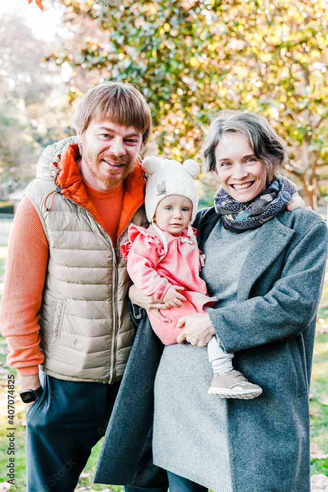 Cheerful parents with cute little baby posing for photo in park together. Blonde mother holding daughter. Family with child standing on grass with fallen maple leaves. Parenthood and weekend concept Cheerful parents with cute little baby posing for photo in park together. Blonde mother holding daughter. Family with child standing on grass with fallen maple leaves. Parenthood and weekend concept