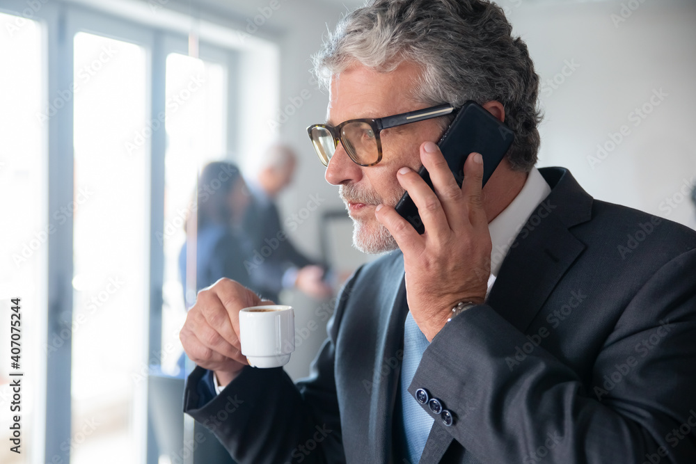 Pensive mature businessman sipping espresso from tiny cup while standing by office glass wall and talking on mobile phone. Copy space. Business and coffee break concept Pensive mature businessman sipping espresso from tiny cup while standing by office glass wall and talking on mobile phone. Copy space. Business and coffee break concept
