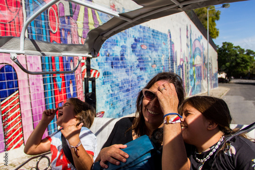 mujer, niño y niña paseando muy alegres en tuk tuk por calles de Portugal, con grafiti de fondo