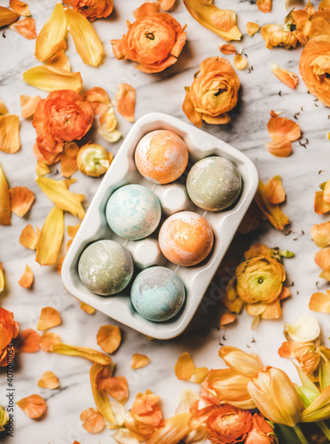 Easter holiday background, texture and wallpaper. Flat-lay of colorful dyed Easter eggs in white ceramic egg holder over blooming orange flowers and white marble background, top view