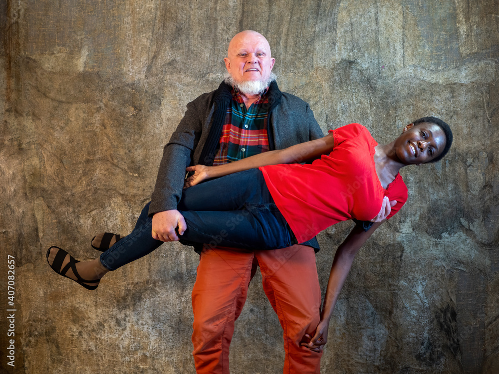 elderly man with gray beard in plaid shirt and red pants lifted beautiful African American girl in his arms and held her. Interracial marriage concept