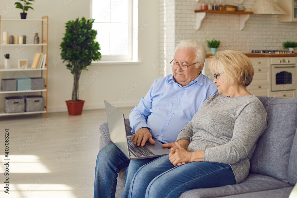 Senior couple spending time together sitting on sofa in living room and reading news or watching movie on laptop. Technology connect and modern elderly lifestyles concept. Senior couple spending time together sitting on sofa in living room and reading news or watching movie on laptop. Technology connect and modern elderly lifestyles concept.