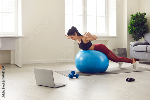 Sporty healthy lifestyle. Slender and sporty woman in sportswear trains at home in the morning with a fitness ball and watches her favorite show on a laptop. Home workout concept.