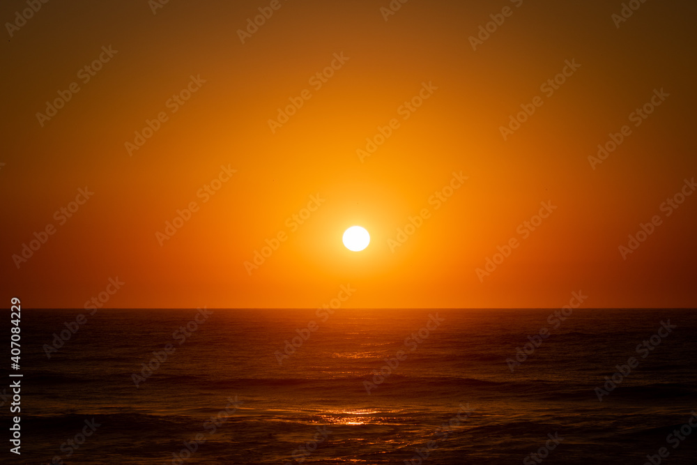 The bright round sun setting above the calm Atlantic Ocean and illuminating the whole clear sky and water in Quiaios Beach, Portugal. Nature background with the sunset and sea