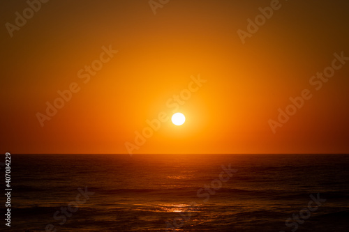 Fototapeta Naklejka Na Ścianę i Meble -  The bright round sun setting above the calm Atlantic Ocean and illuminating the whole clear sky and water in Quiaios Beach, Portugal. Nature background with the sunset and sea