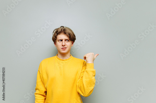 Displeased young man in yellow clothes shows finger to the side on looking with suspicious face on copy space, isolated on gray background.