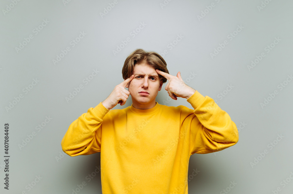 Serious guy in yellow clothes looks at the camera and points his fingers at his temple on a background of a gray wall. Copy space Serious guy in yellow clothes looks at the camera and points his fingers at his temple on a background of a gray wall. Copy space