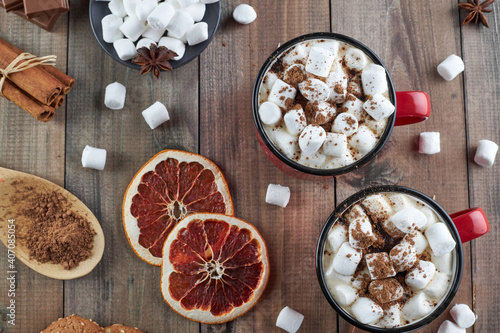 Two red cups of hot chocolate with marshmallow sprinkled with cocoa powder on a wooden table with slices of dried grapefruit and chocolate pieces. Winter hot drink with spices. Top view.